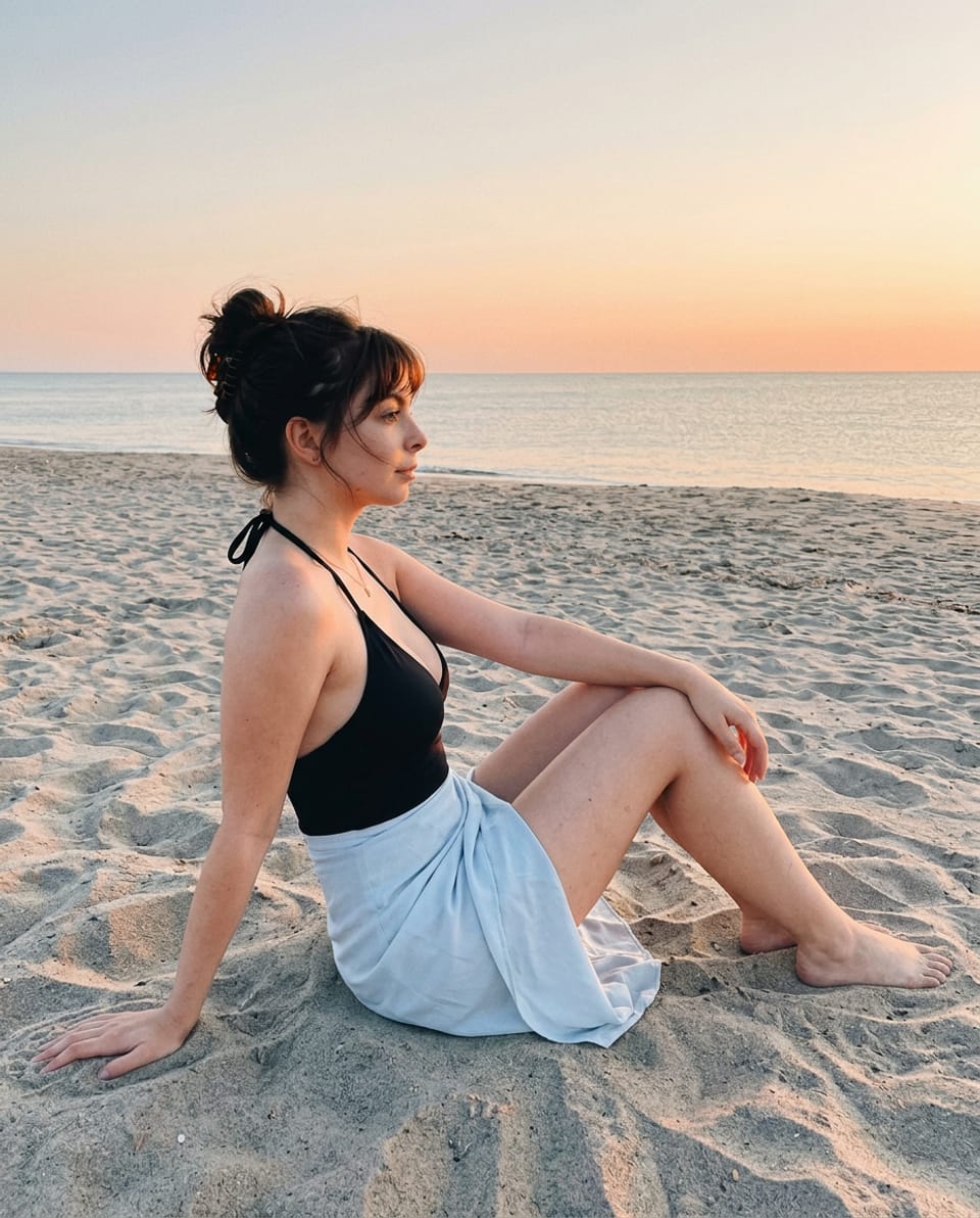 Woman in a black halter swimsuit and pale-blue wrap sitting on dry sand, one palm behind her hip and the other hand over a bent knee at sunset.